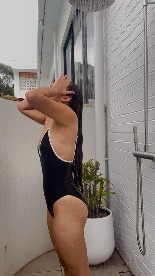 Person in a black swimsuit standing under a shower with a plant nearby.