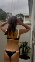 Person wearing a black and white bikini standing under a shower with a plant in the background.